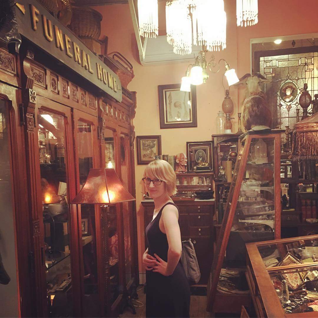 A woman stands next to a large wooden display case filled with antiques.